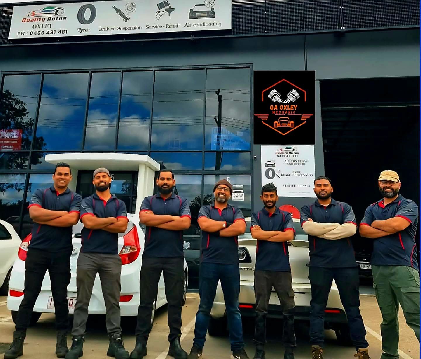 Seven men in matching uniforms stand in front of an auto repair shop with arms crossed.