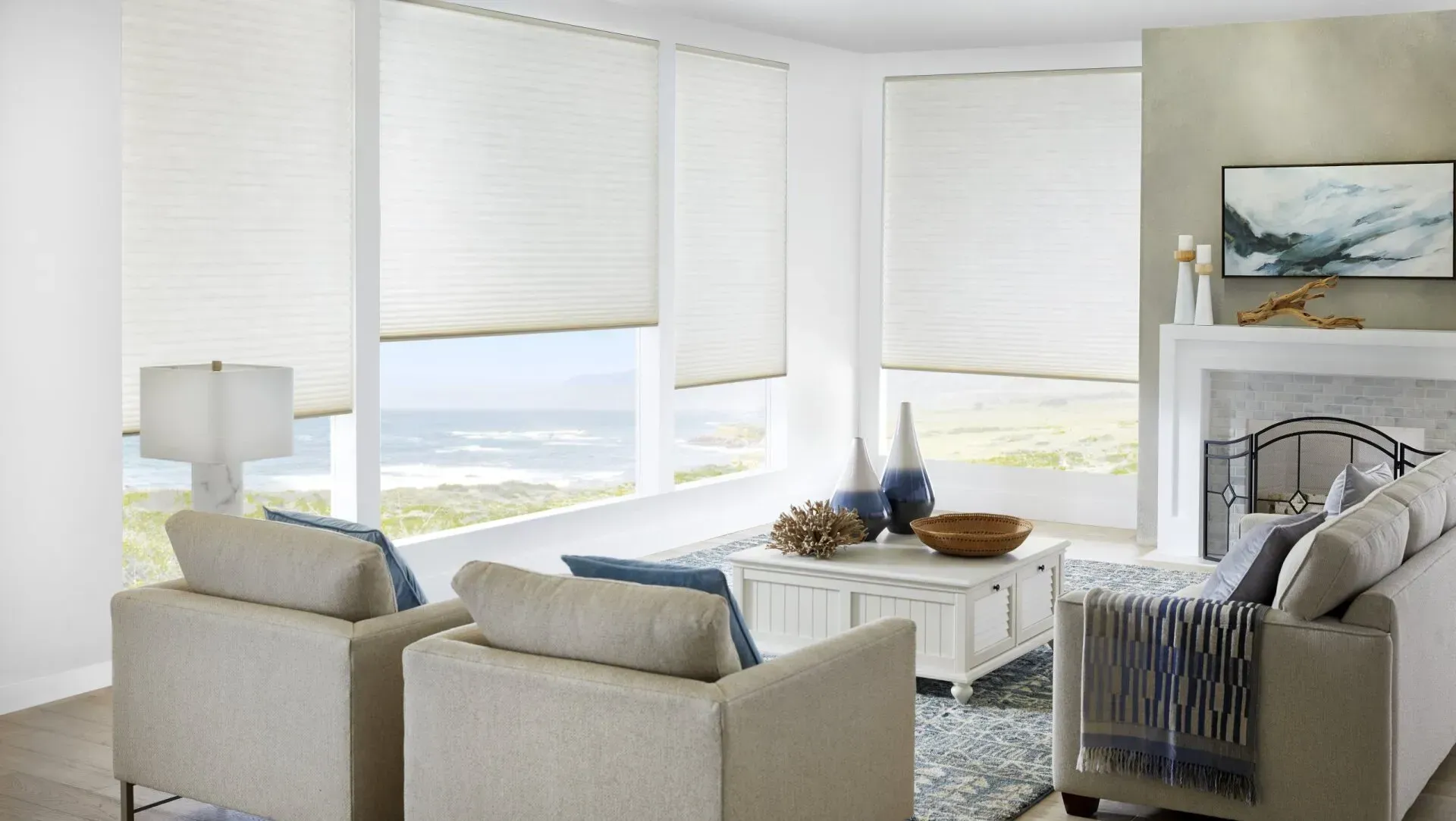 A living room with white honeycomb shades, ocean view, two armchairs, a sofa, fireplace, and a coffee table.