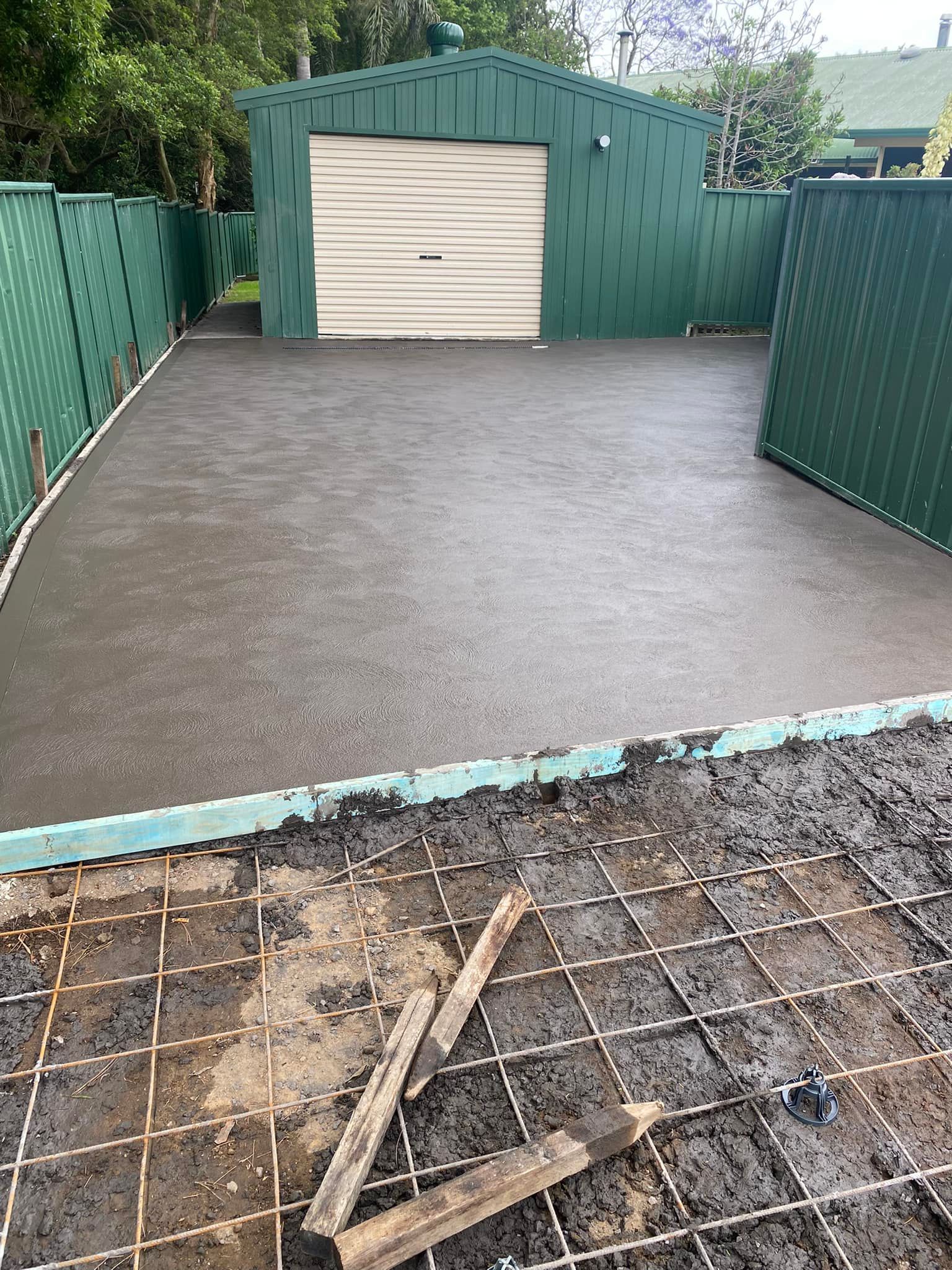 Concrete patio area enclosed by green fences, with a small shed and unfinished ground in the foreground. — BJ's Concreting in Huskisson, NSW