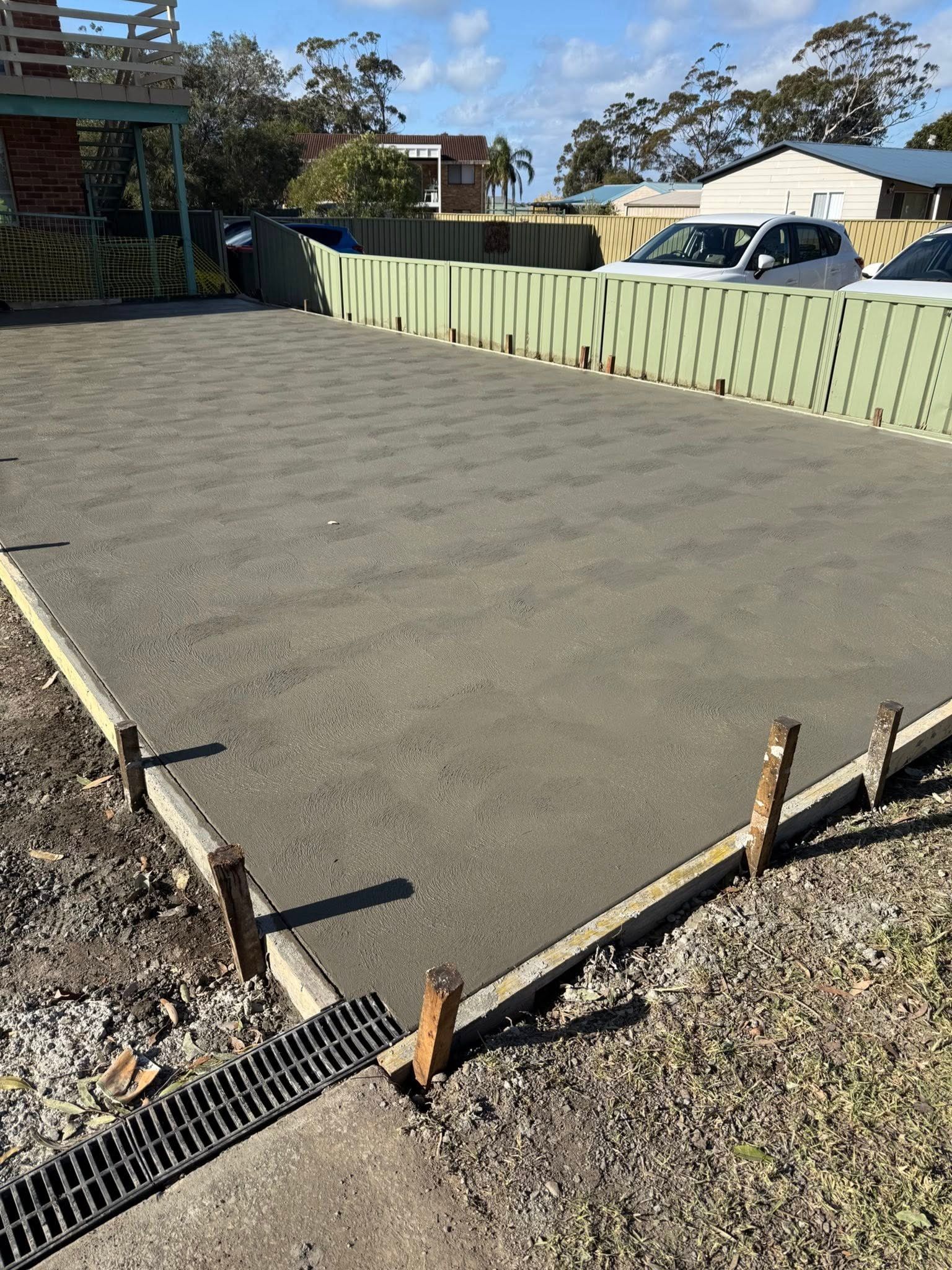 Freshly poured concrete slab under construction beside a green fence and gravel driveway — BJ's Concreting in Huskisson, NSW