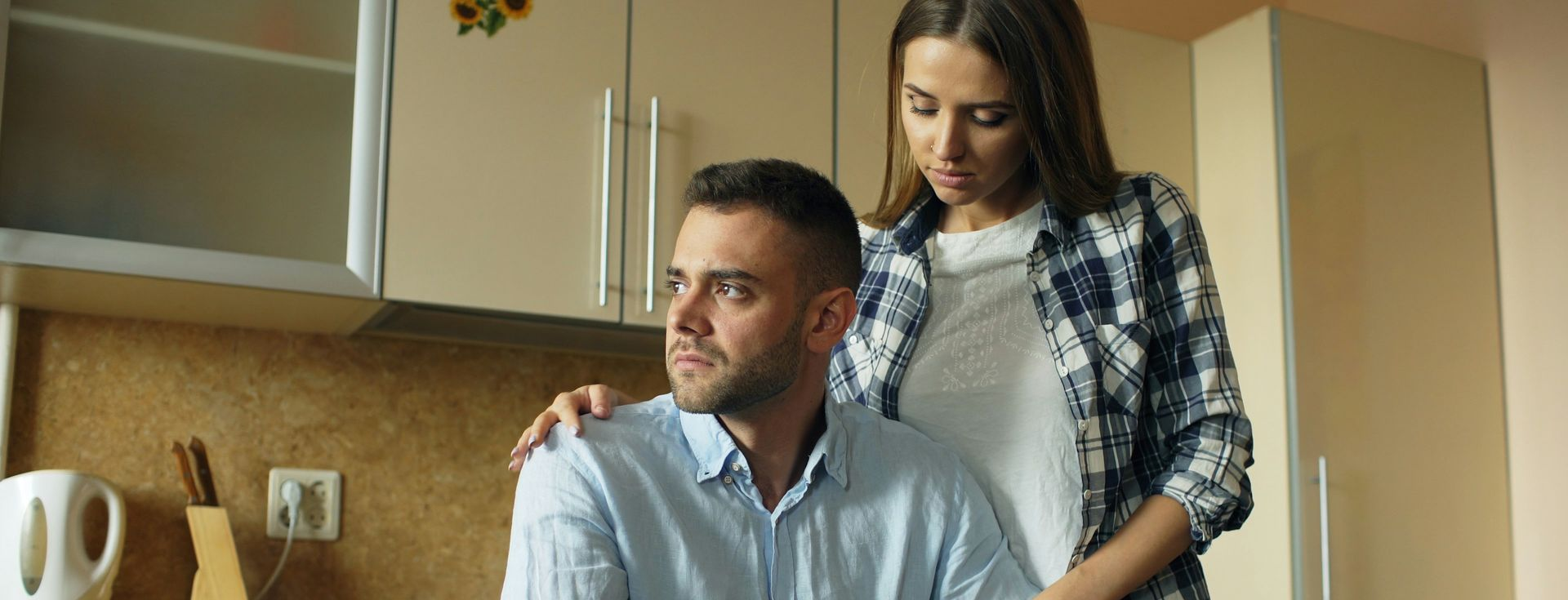 Man kijkt boos weg. Vrouw staat achter hem met hand op schouder.