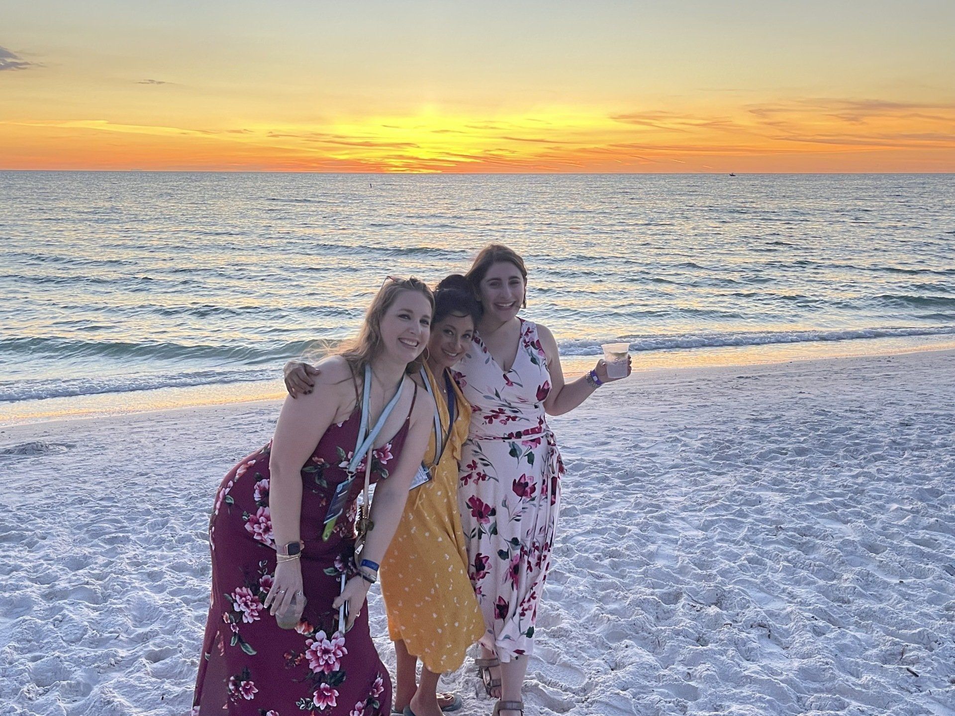 Three women are posing for a picture on the beach at sunset.
