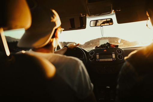 Two people in a car driving towards the sun. The driver wears a baseball cap. Interior view.