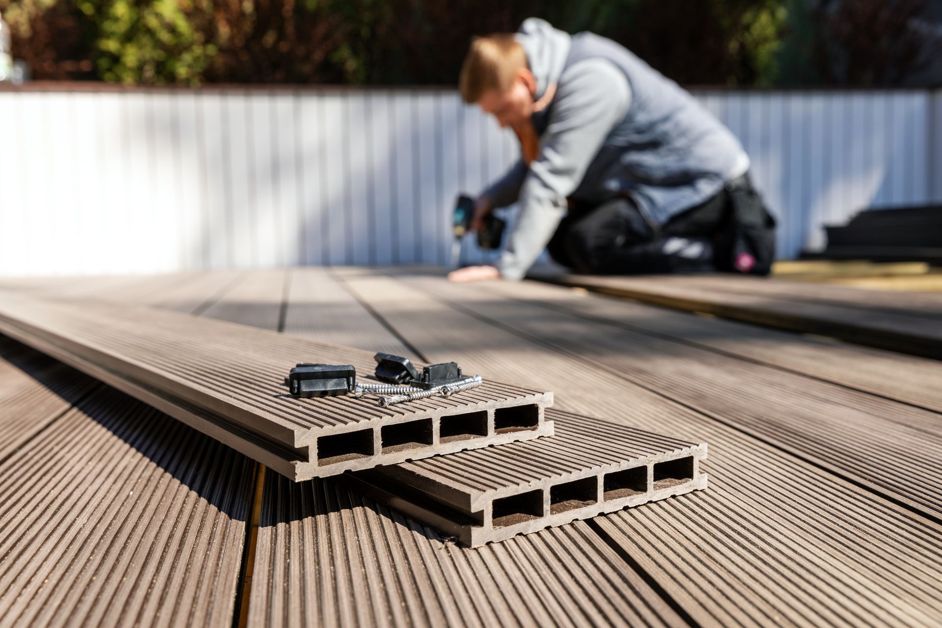 Person installing composite decking with a drill outdoors; close-up on brown boards and fasteners. Person installing composite decking with a drill outdoors; close-up on brown boards and fasteners.