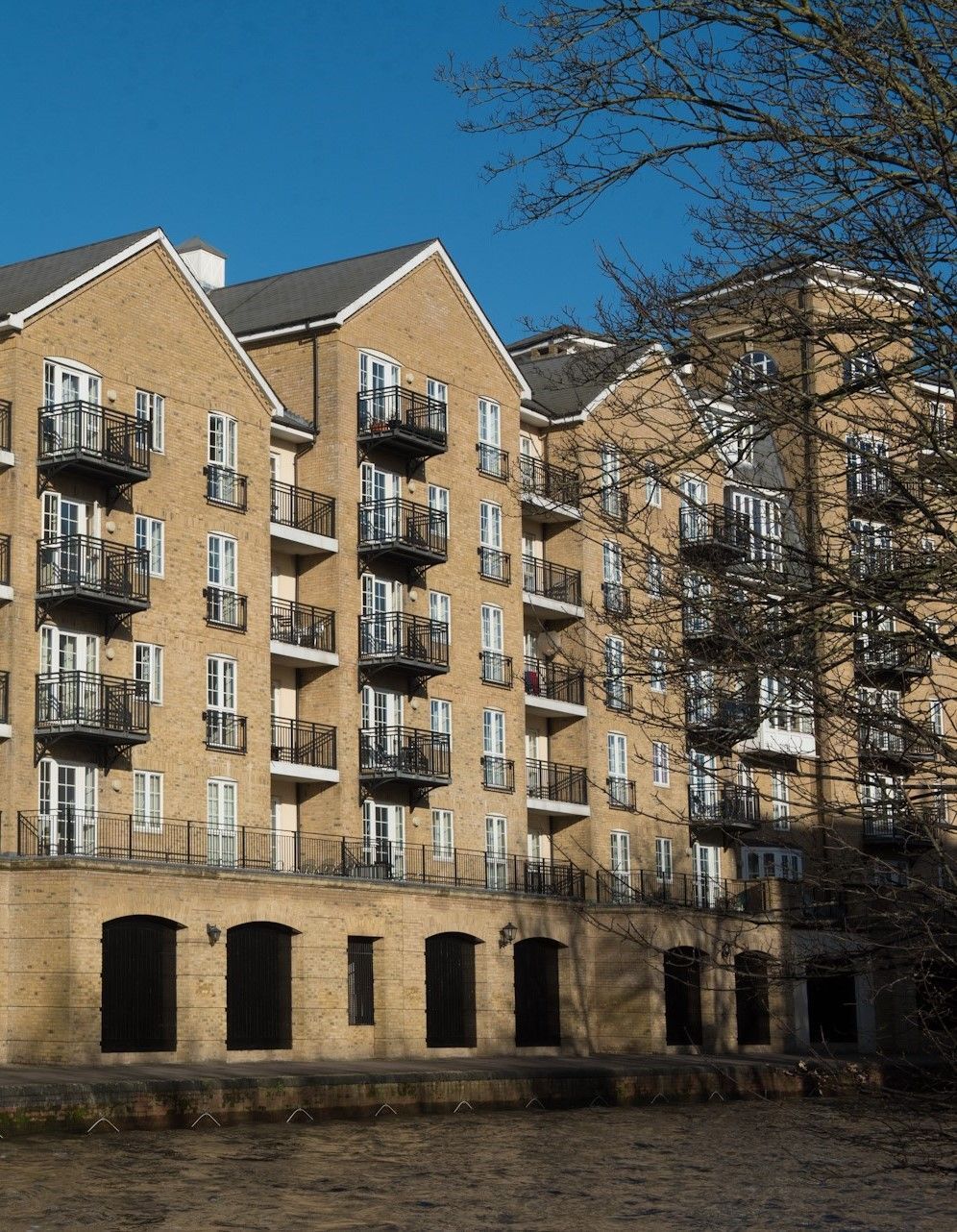 Yellow-brick riverside house with balconies, beside water, under a clear blue sky.