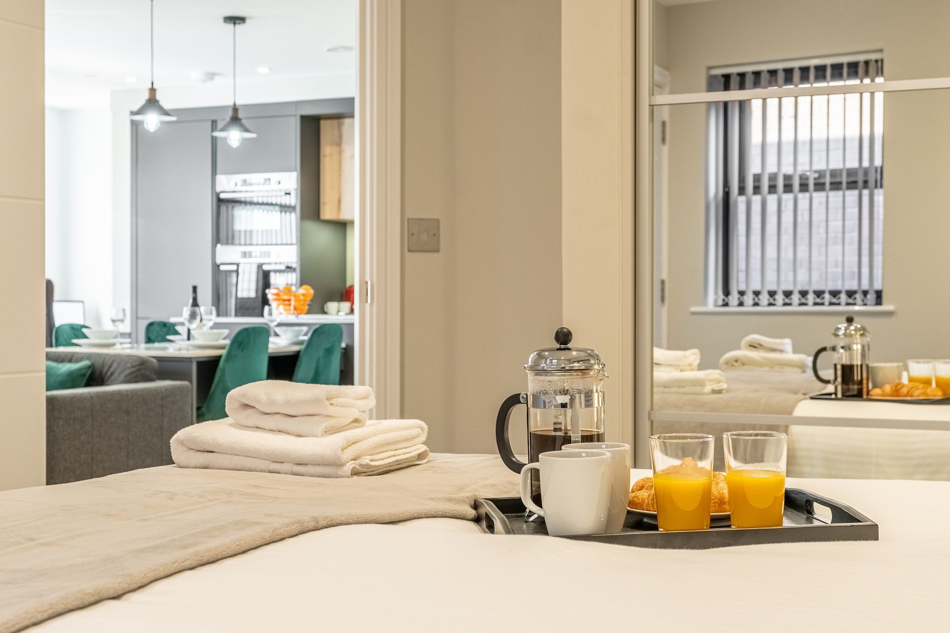 Tray with coffee, orange juice, and pastries on bed; folded towels and dining area in background.