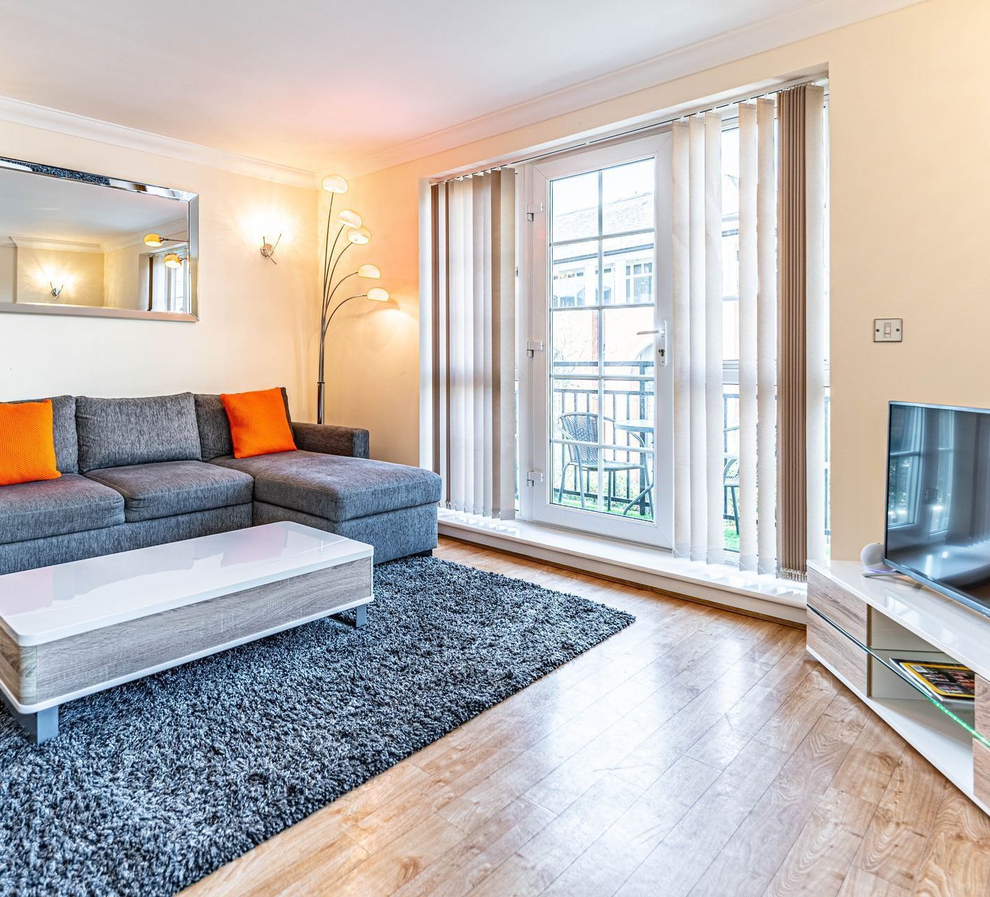 Modern living room with gray sofa, orange cushions, coffee table, rug, TV, and large window.
