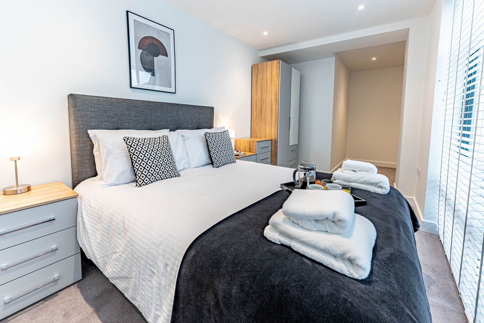 A modern bedroom with a neatly made double bed, two nightstands, a wardrobe, and towels and a coffee tray placed on the bed. The room is illuminated by natural light from a window with blinds.