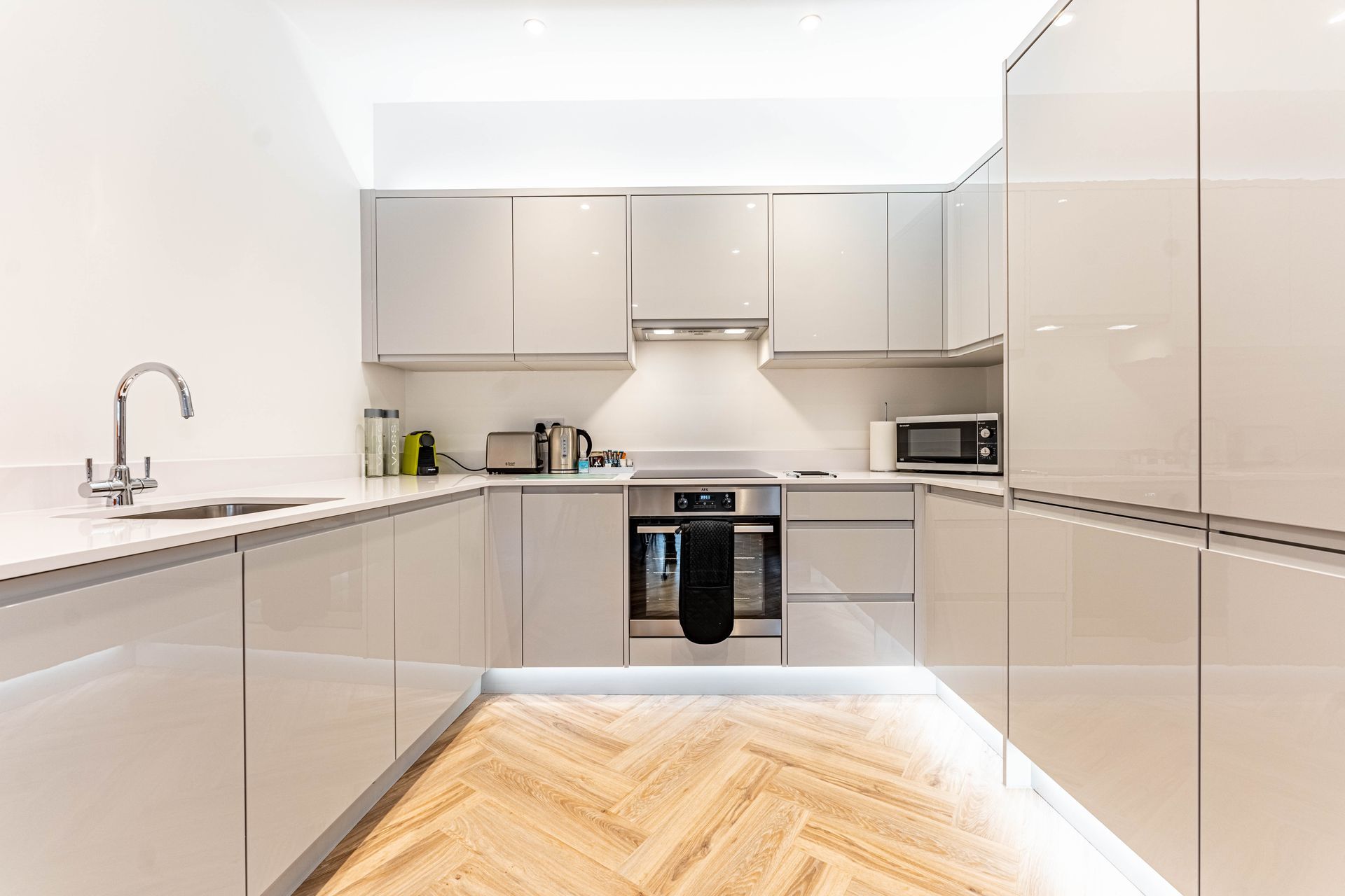 Serviced Apartment in Reading - A modern kitchen featuring sleek light gray cabinets, a built-in stove with a black towel, a sink, countertop appliances like a toaster, and a microwave. The floor is a herringbone wood pattern.