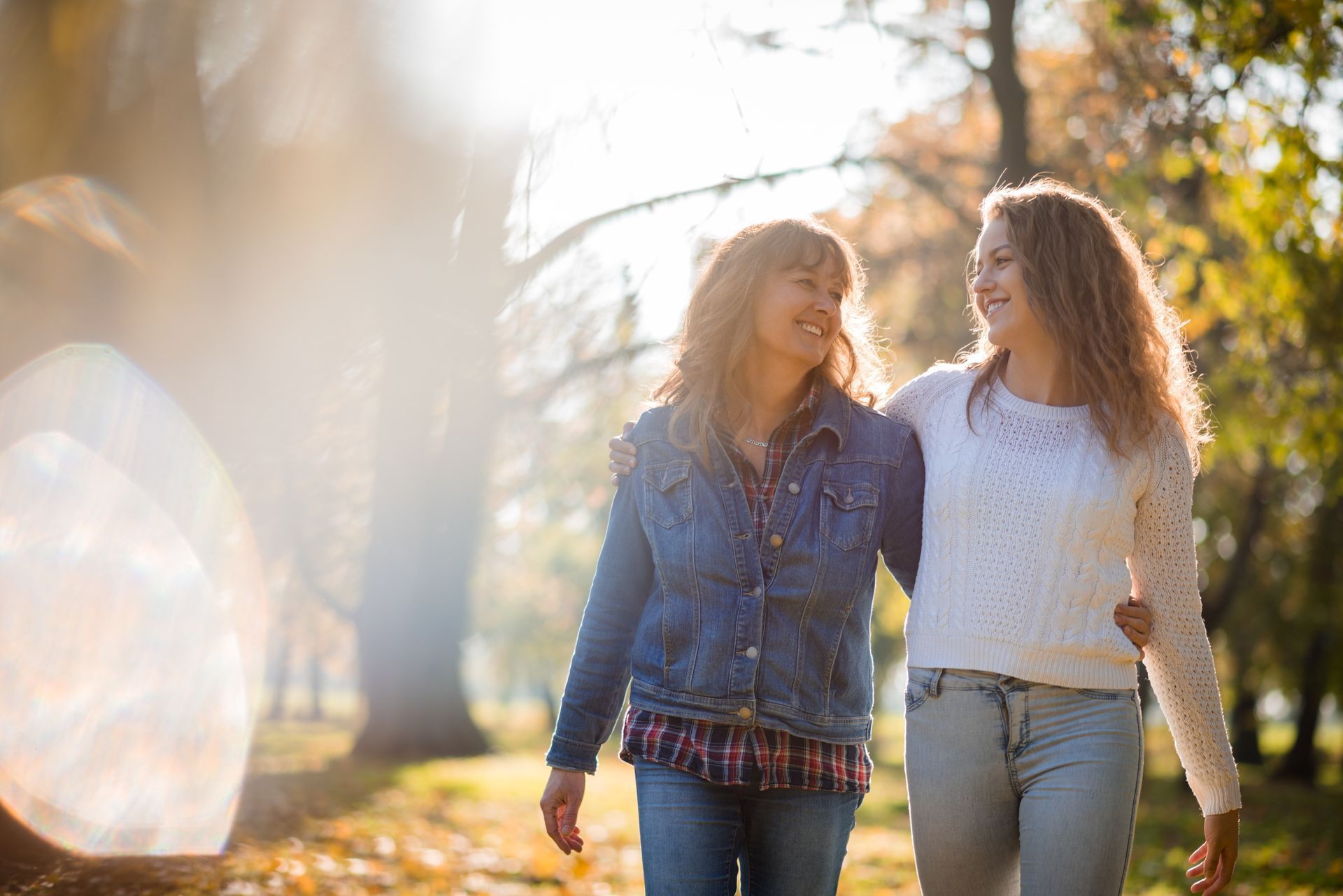 A mother and daughter walk together in a sunlit park, smiling and enjoying a heartfelt conversation during an autumn day.