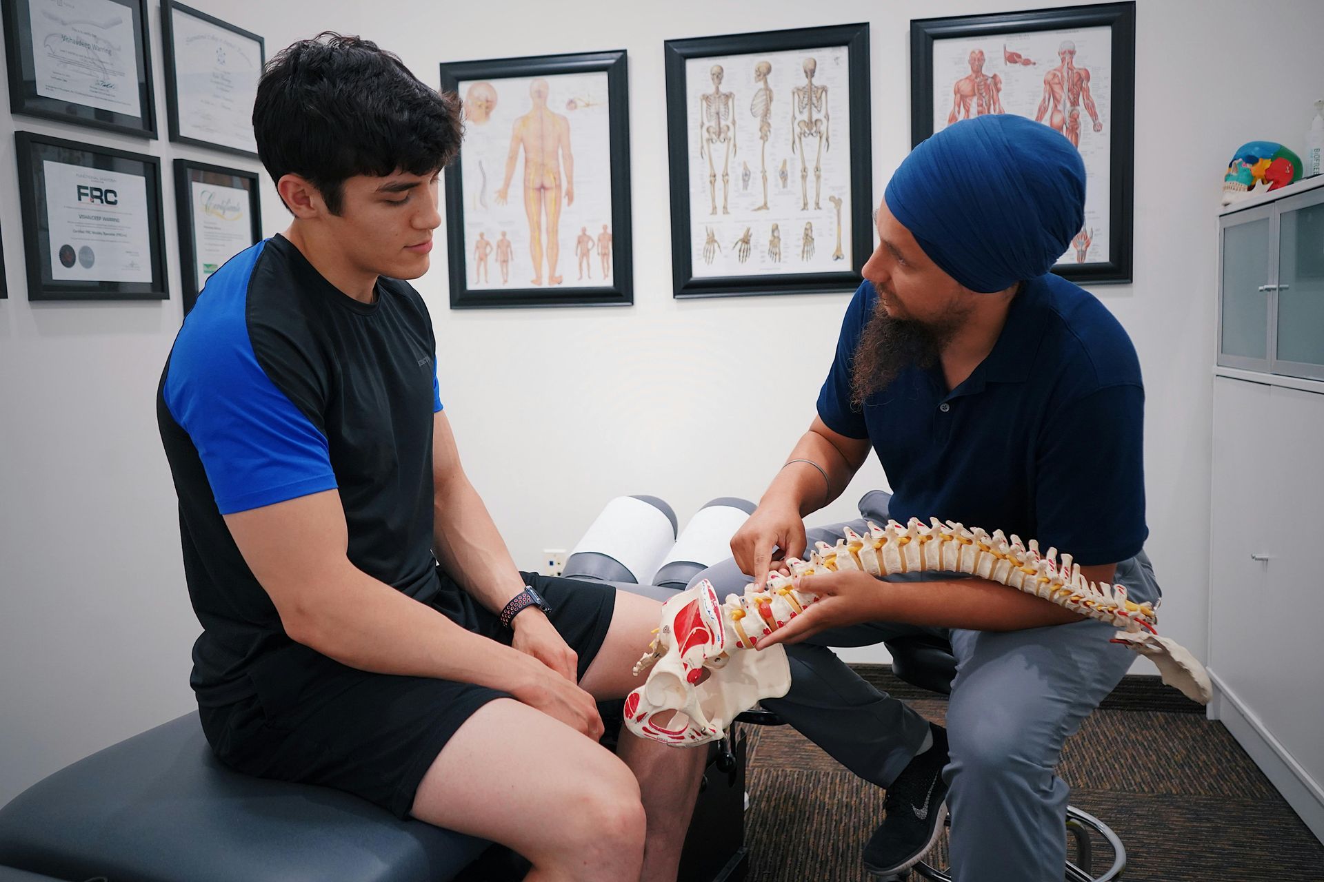 Patient in a clinic receives a spine assessment from a practitioner beside anatomical posters.