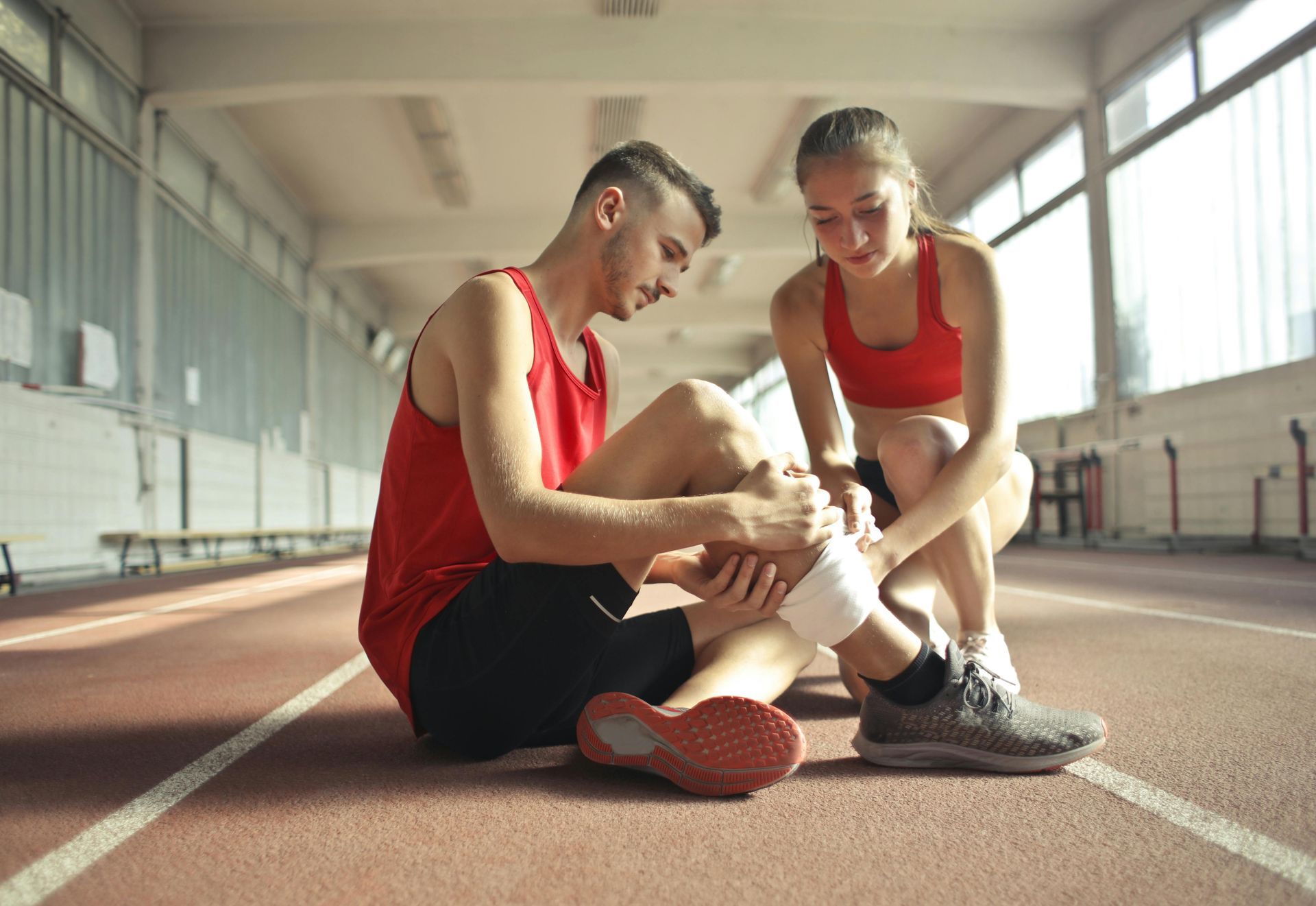 Two athletes in red jerseys sit on an indoor track, one tying the other’s shoe
