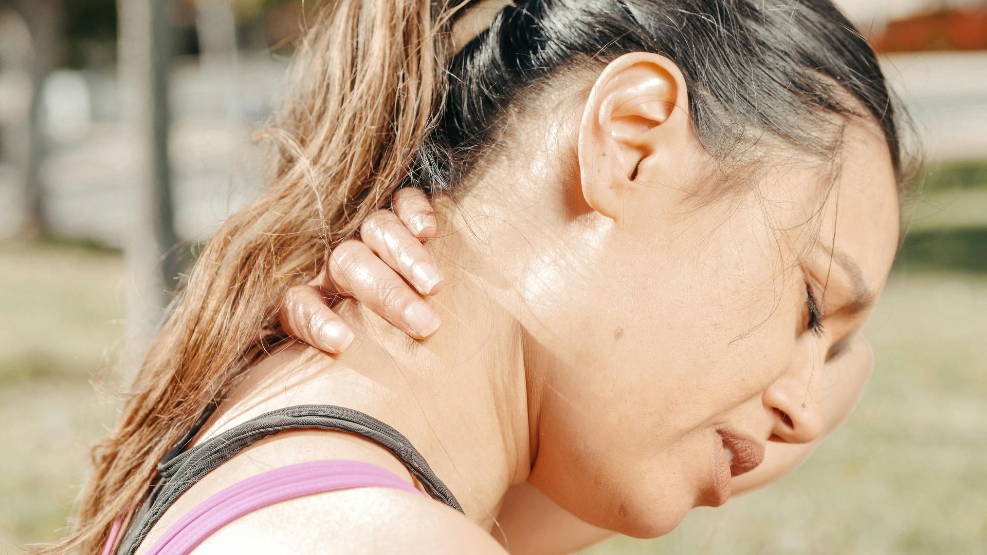 Person holding neck in pain outdoors, with hand on shoulder and eyes closed