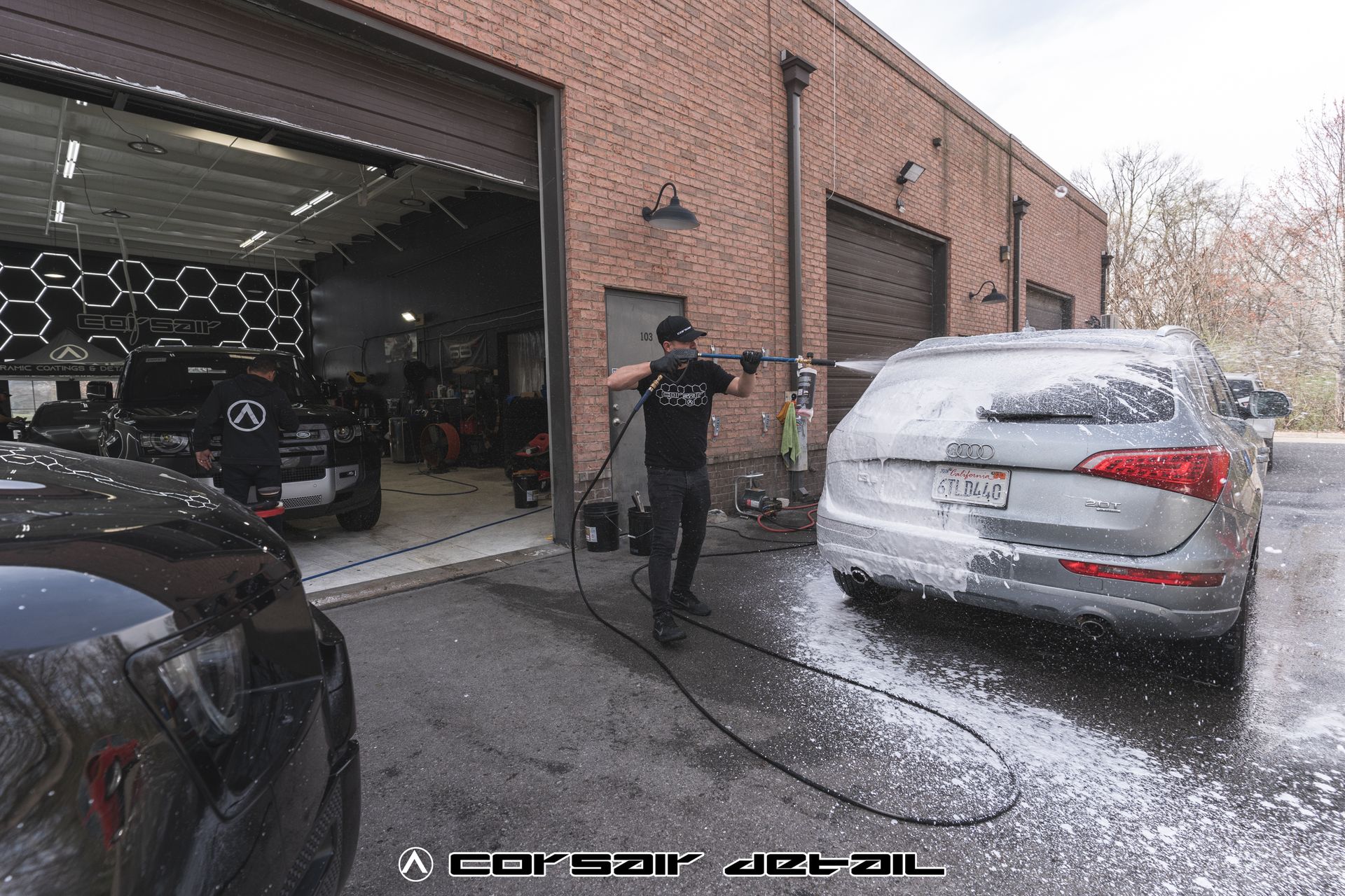 A man is washing a car with a high pressure washer in front of a garage.