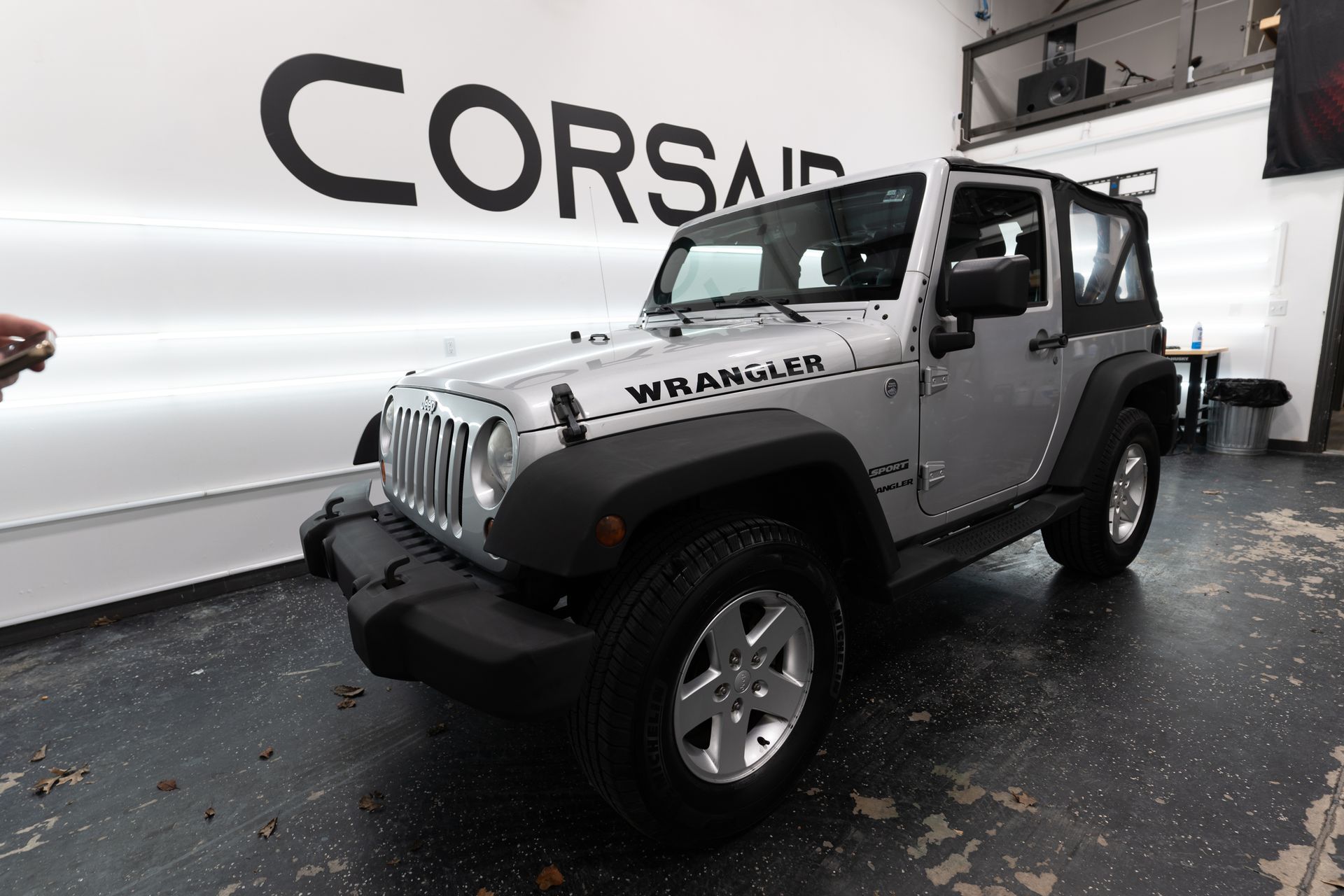 A silver jeep wrangler is parked in front of a wall that says corsair.