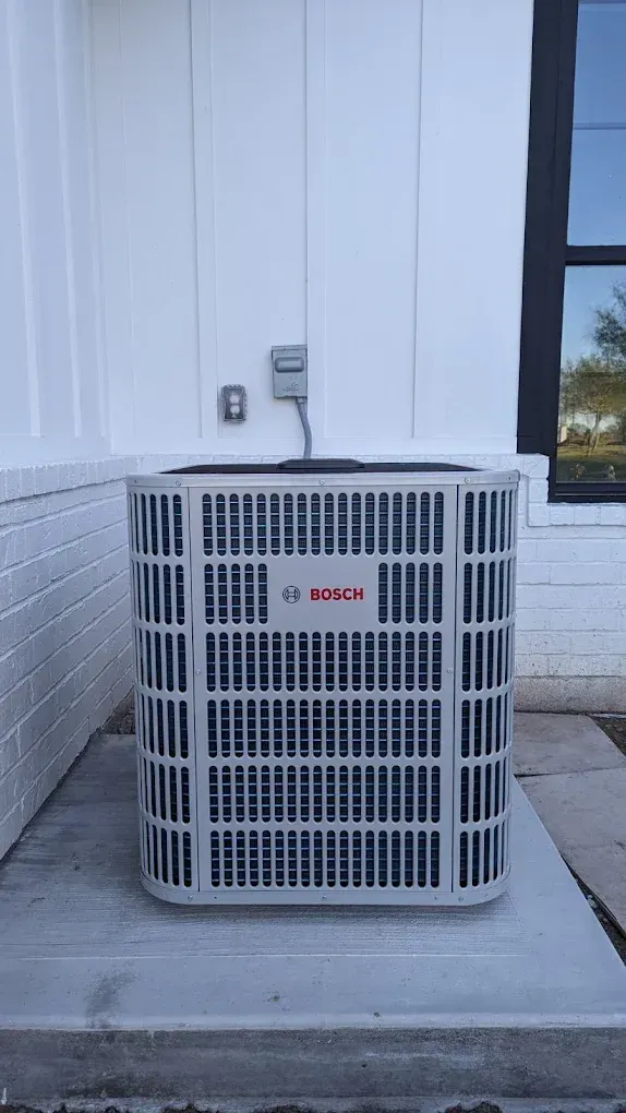 Bosch air conditioning unit on concrete pad, mounted on exterior white wall. Electrical box above.