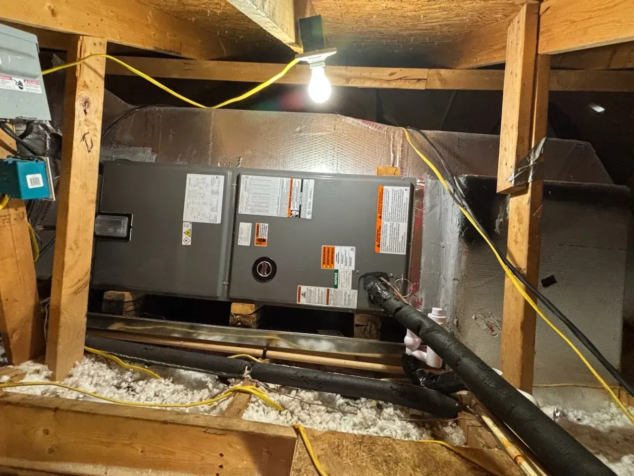 HVAC unit in an attic with wooden beams, insulation, and a bare lightbulb.