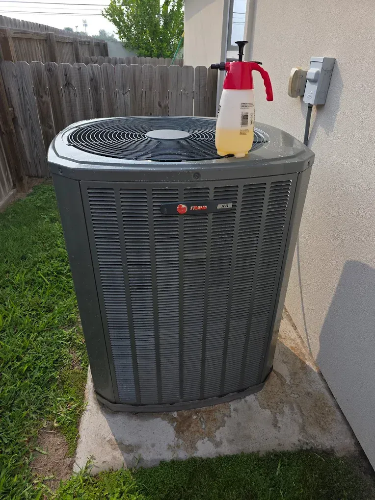 Gray Trane air conditioner unit with a red and white sprayer bottle on top, next to a wall.