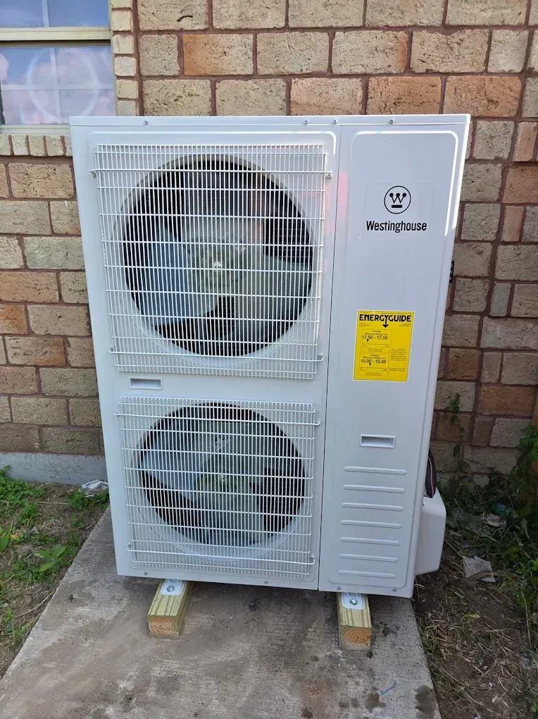 Outdoor heat pump unit, white with two fan compartments, set on wooden blocks next to a brick wall.