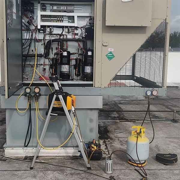 HVAC unit on a rooftop being serviced. A technician is using gauges and a yellow refrigerant tank.