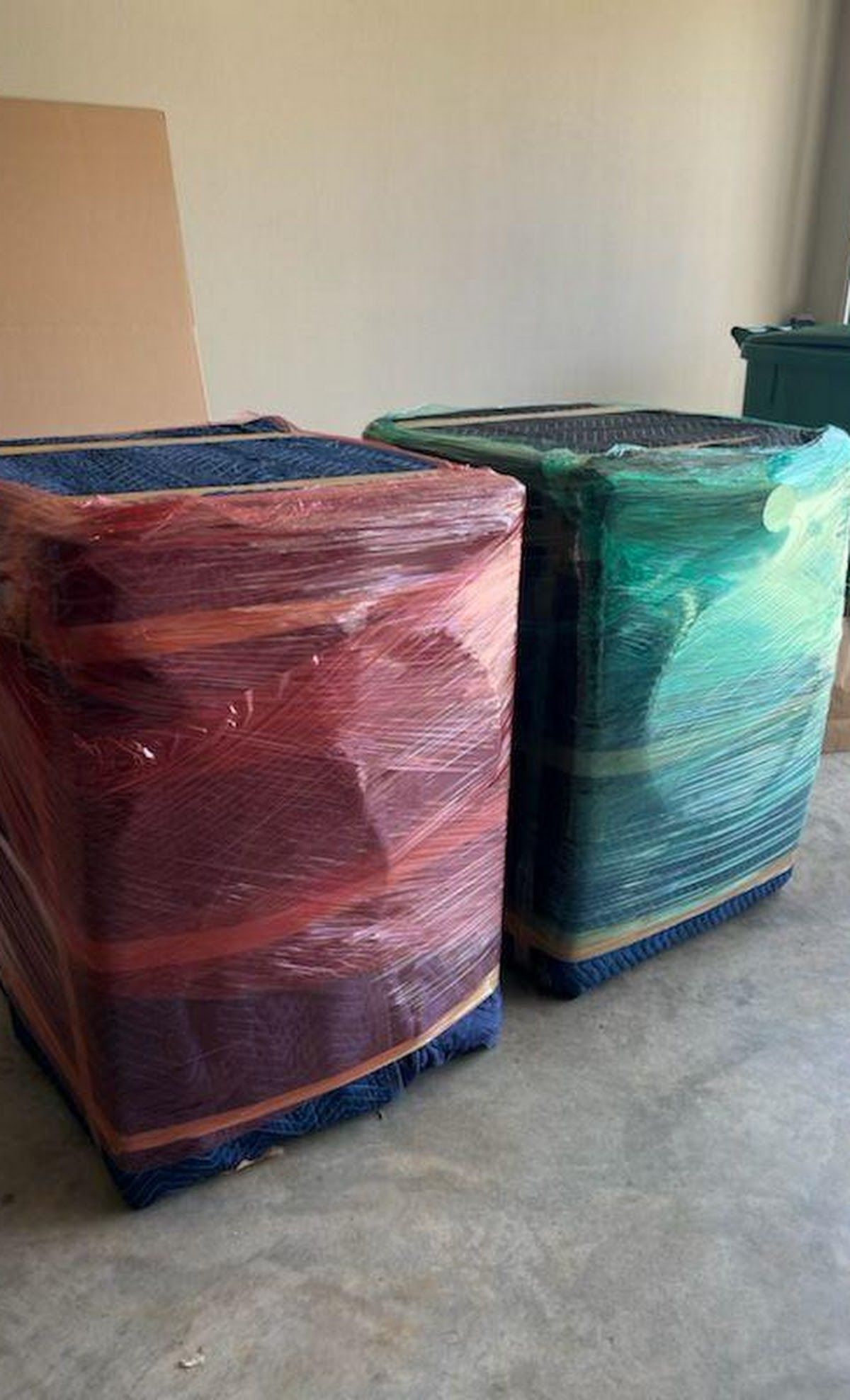 Two upholstered ottomans wrapped in protective plastic, one red and one green, on a concrete floor.