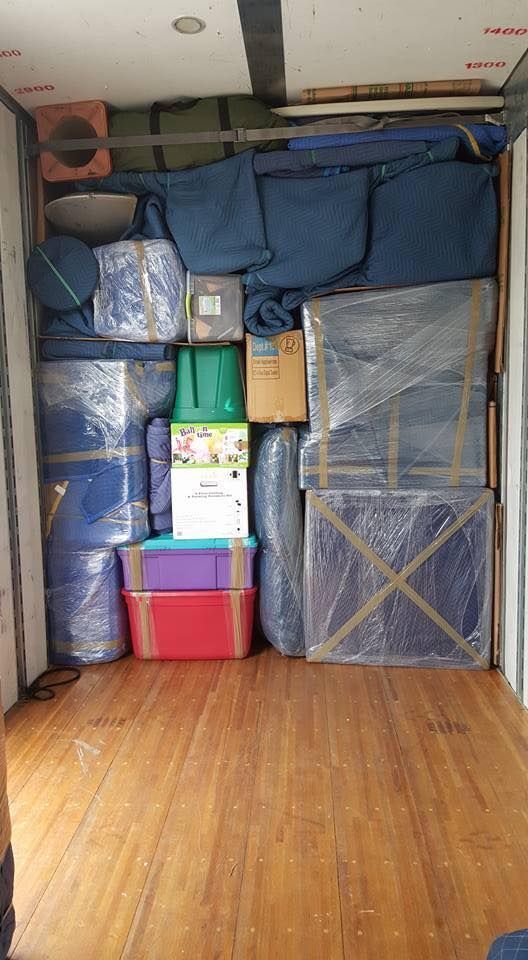 Inside of a moving truck packed with boxes wrapped in plastic and blankets, on a wooden floor.