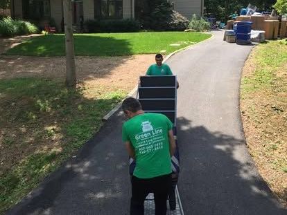 Two movers in green shirts transport a black shelving unit on a dolly down a driveway, towards a house.