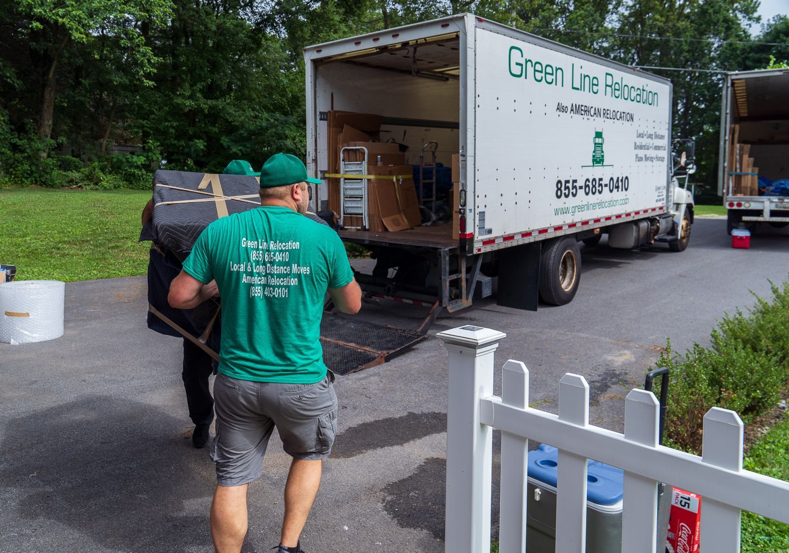 Two movers in green shirts transport a black shelving unit on a dolly down a driveway, towards a house.