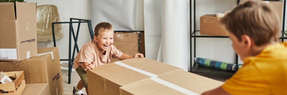 Two children helping to move a large cardboard box in a room with packed boxes. One child smiles, pushing the box.