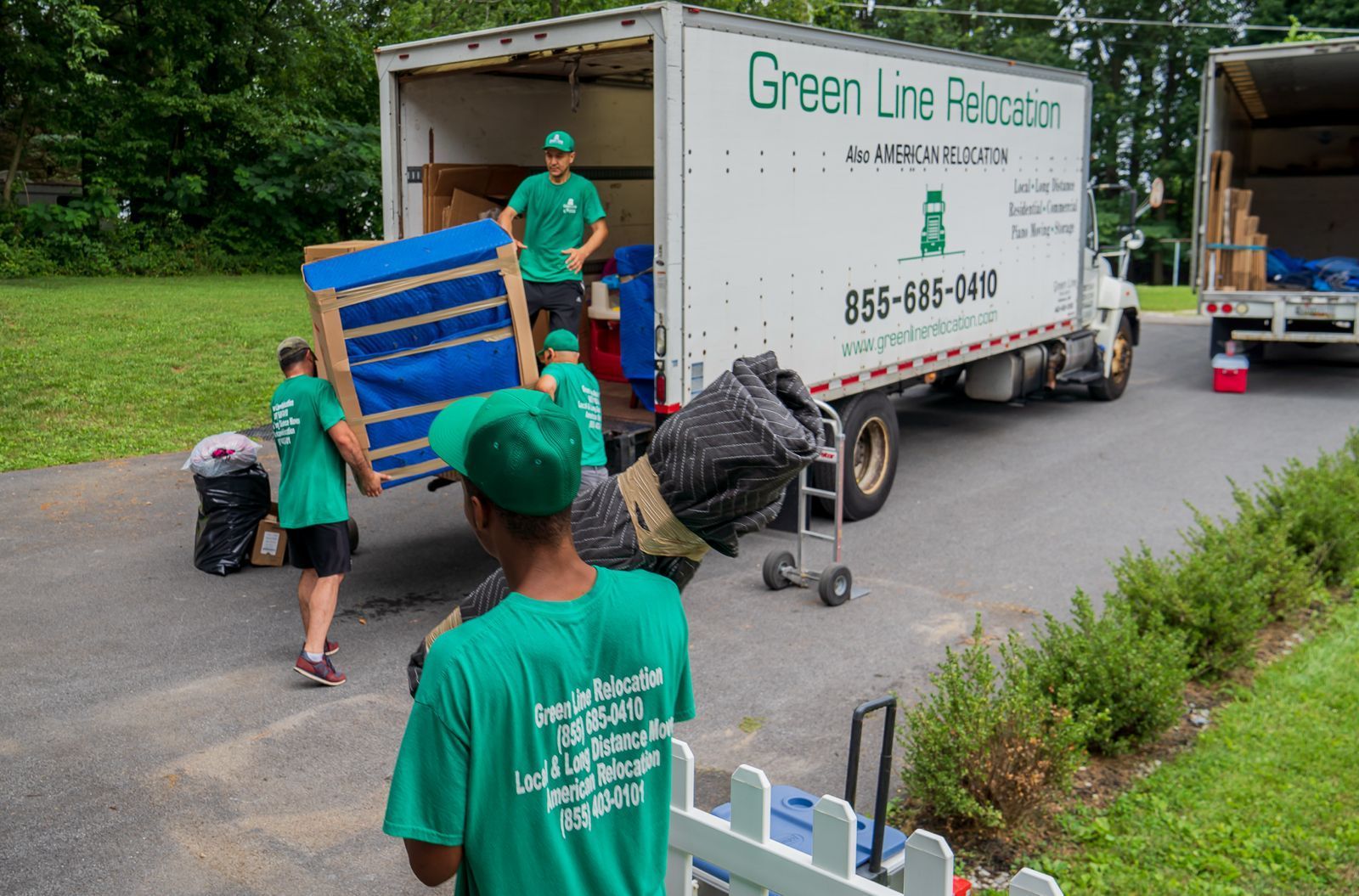 Movers in matching green uniforms load furniture and equipment into a Green Line Relocation truck parked on a paved driveway.