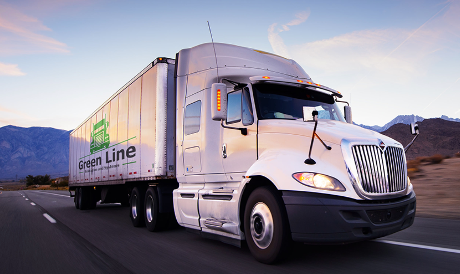 Semi-truck with a white cab and green trailer on a highway, mountains in the background, under a partly cloudy sky.
