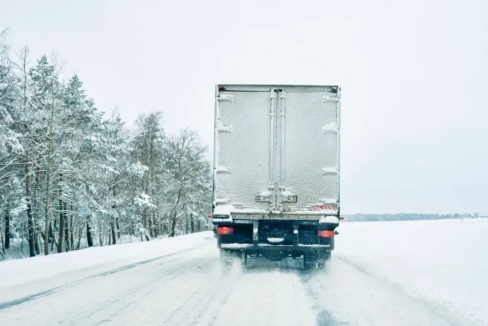 Truck driving on a snow-covered road with trees in the background.