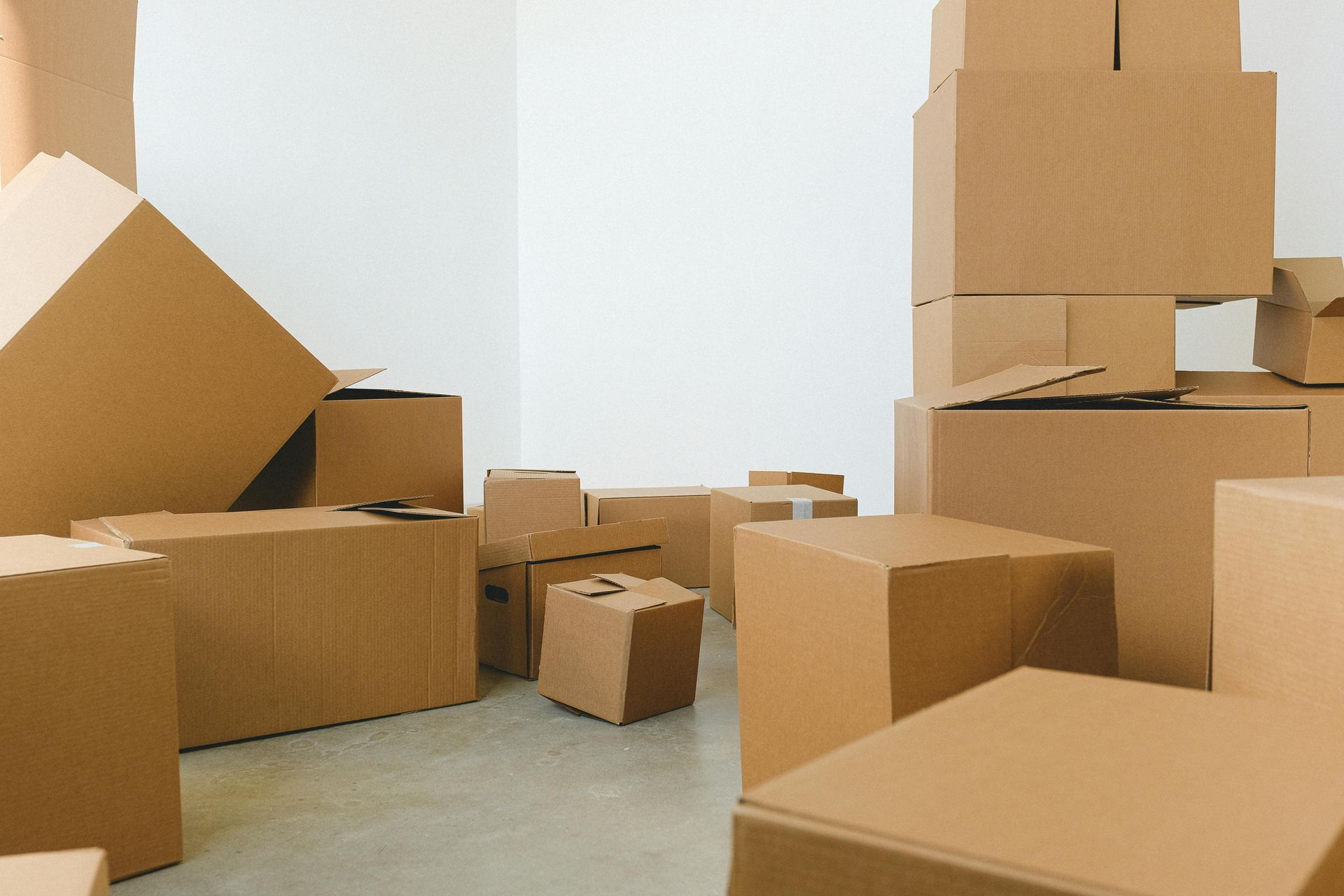 A collection of stacked and scattered cardboard moving boxes of various sizes on a plain floor against a white wall.