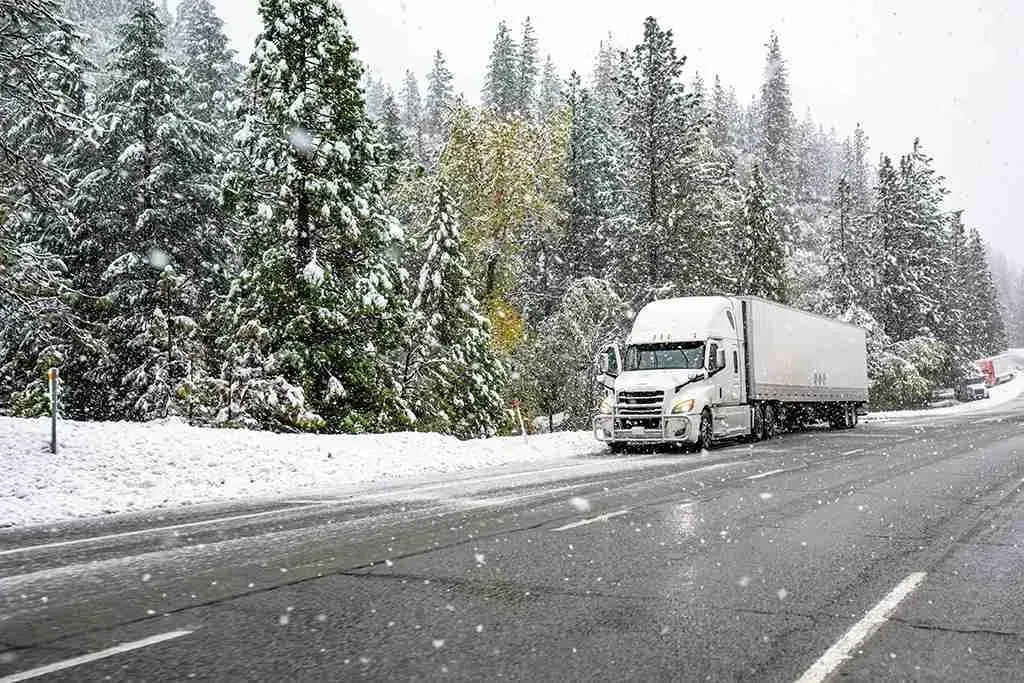 Semi-truck driving on a snowy highway, flanked by snow-covered evergreen trees.