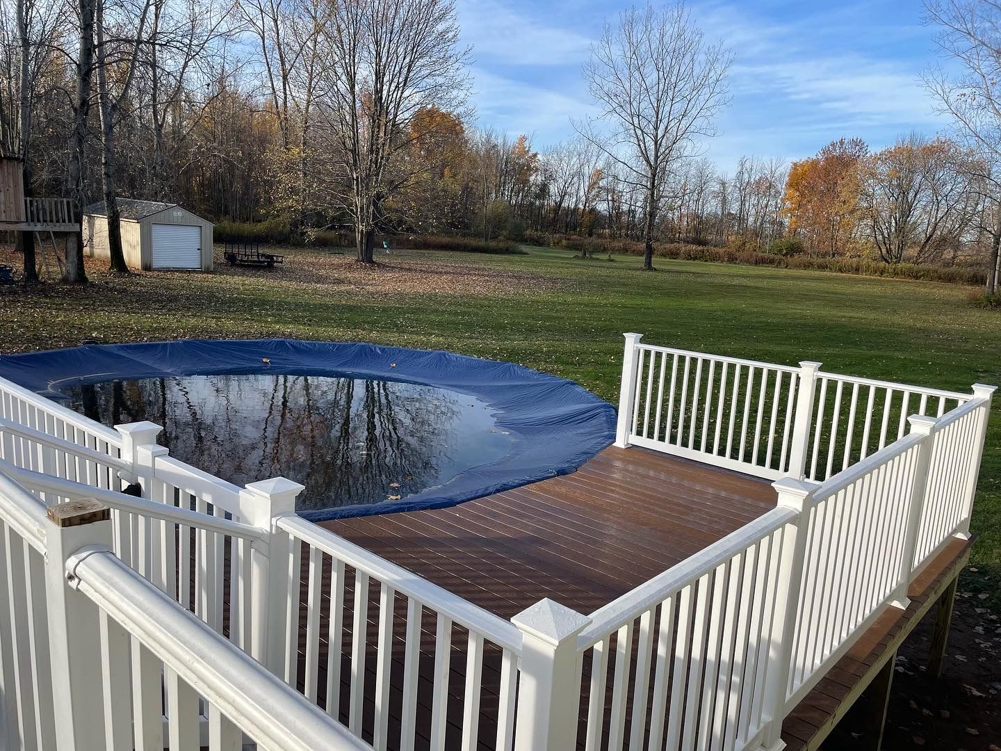 Deck with white railing overlooking a pool covered with blue tarp, grassy backyard, trees.