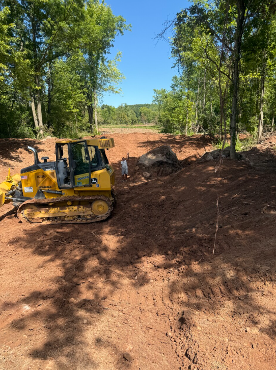 A yellow bulldozer on red dirt path surrounded by trees under a bright blue sky.