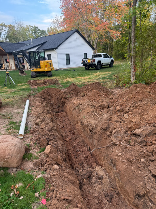 Dug trench with exposed dirt, white house, yellow excavator, white truck, green grass, fall foliage.