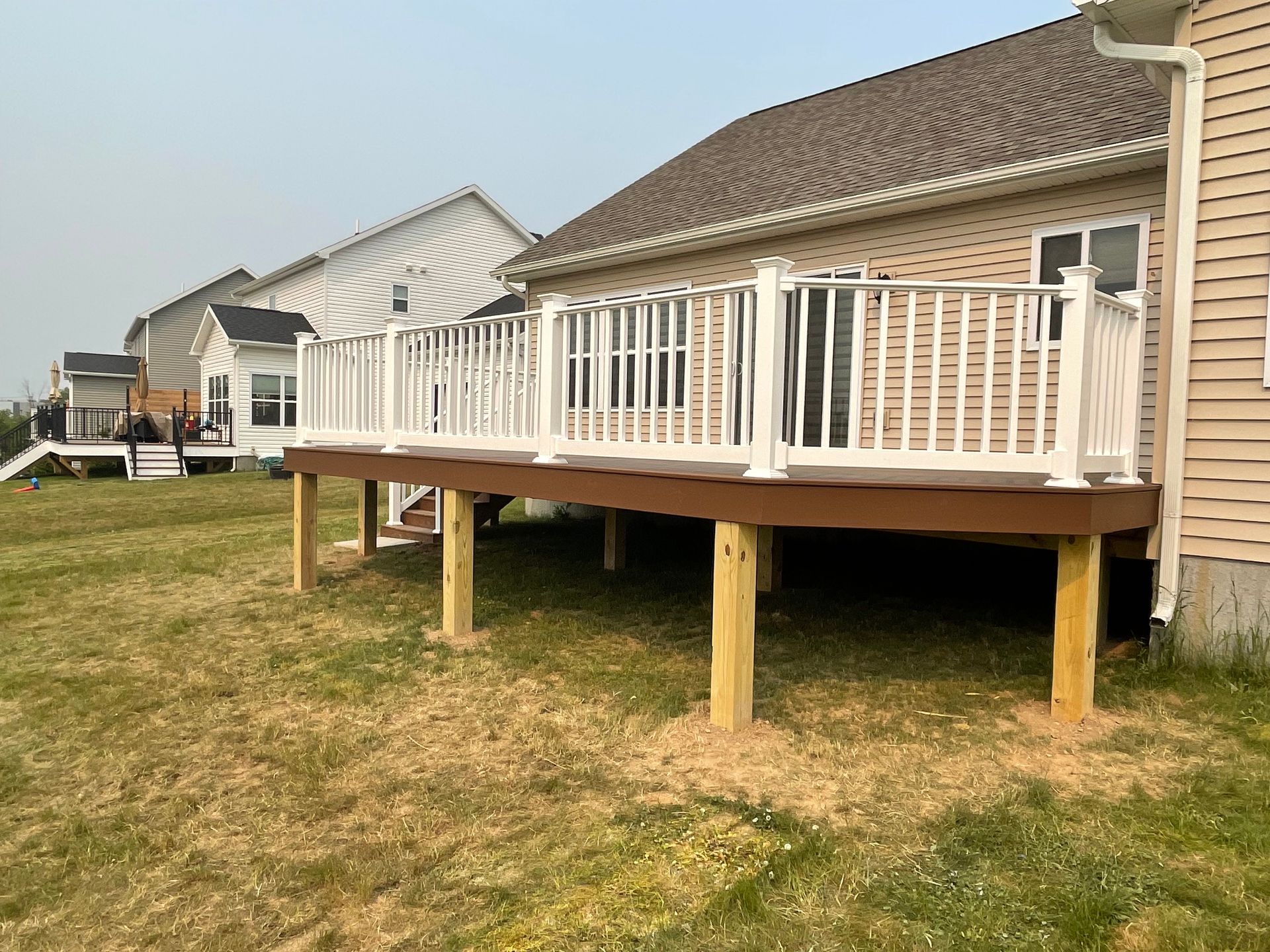A wooden deck with white railing attached to a beige house, standing on wooden posts in a grassy yard.