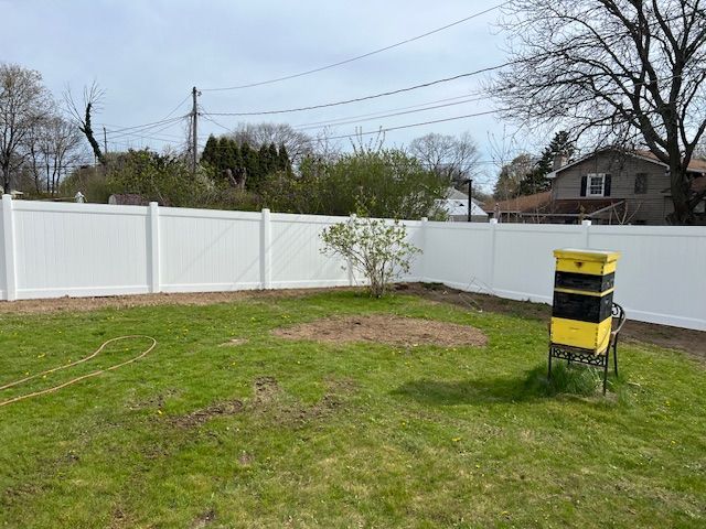 White vinyl fence encloses a grassy backyard with a beehive and a small tree.