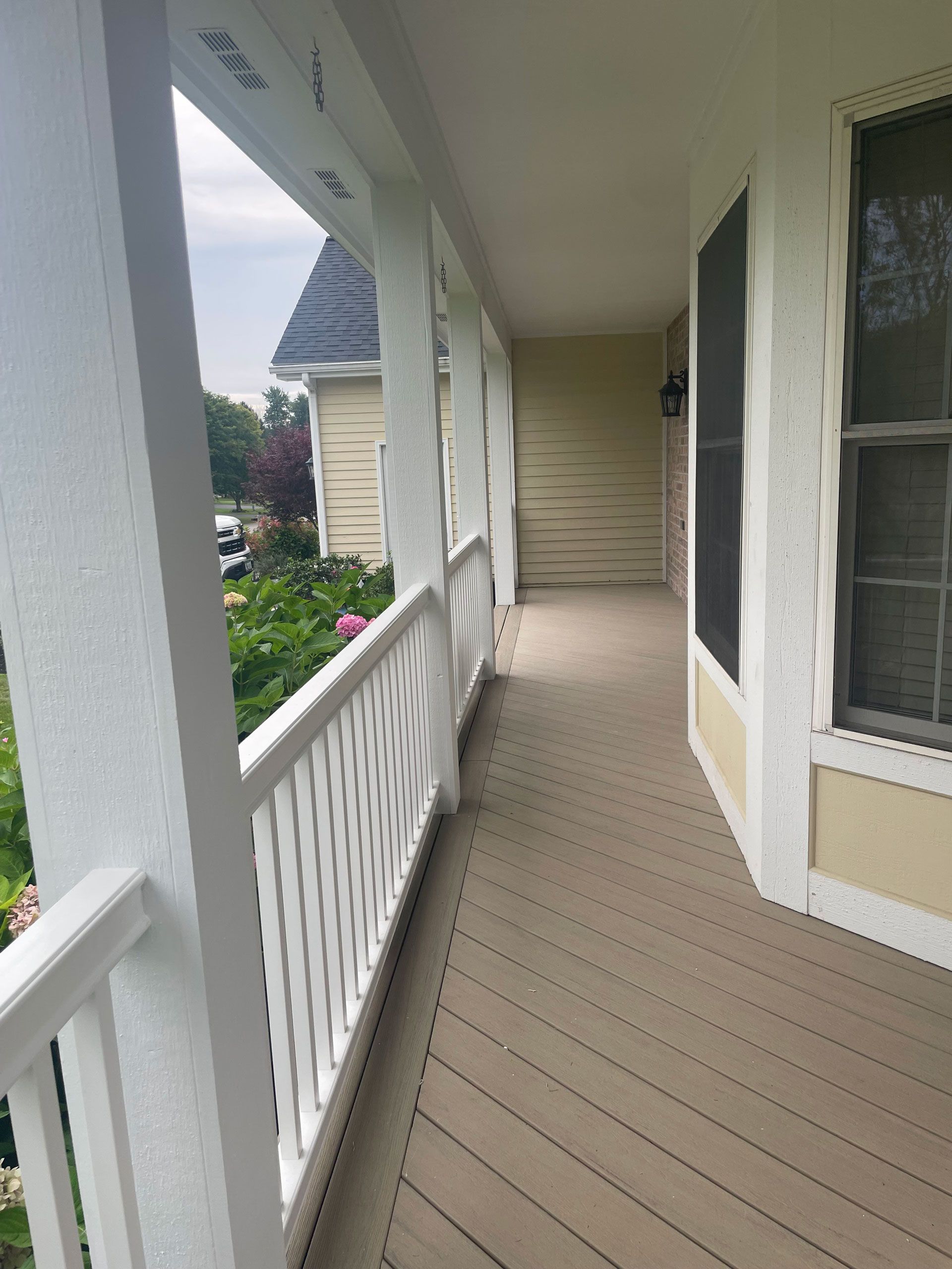 Covered porch with white railings, tan floor, and a view of greenery.