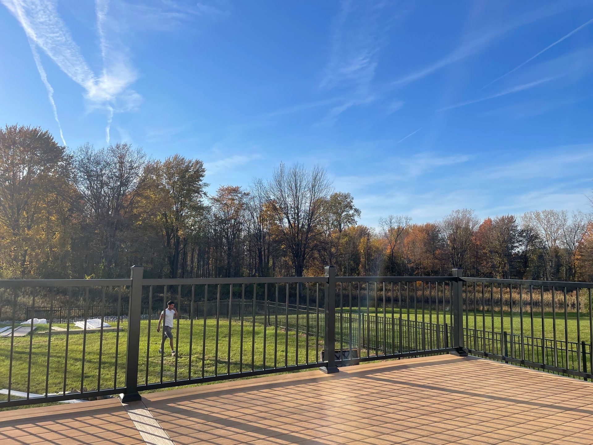 A brick patio with a black fence overlooks a grassy yard and trees under a blue sky.