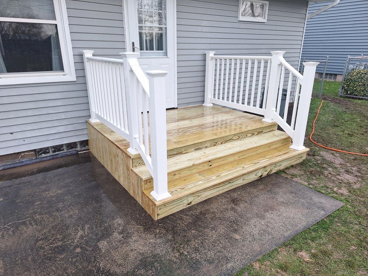 Wooden porch with white railings, steps leading to a door. Gray siding, wet concrete.