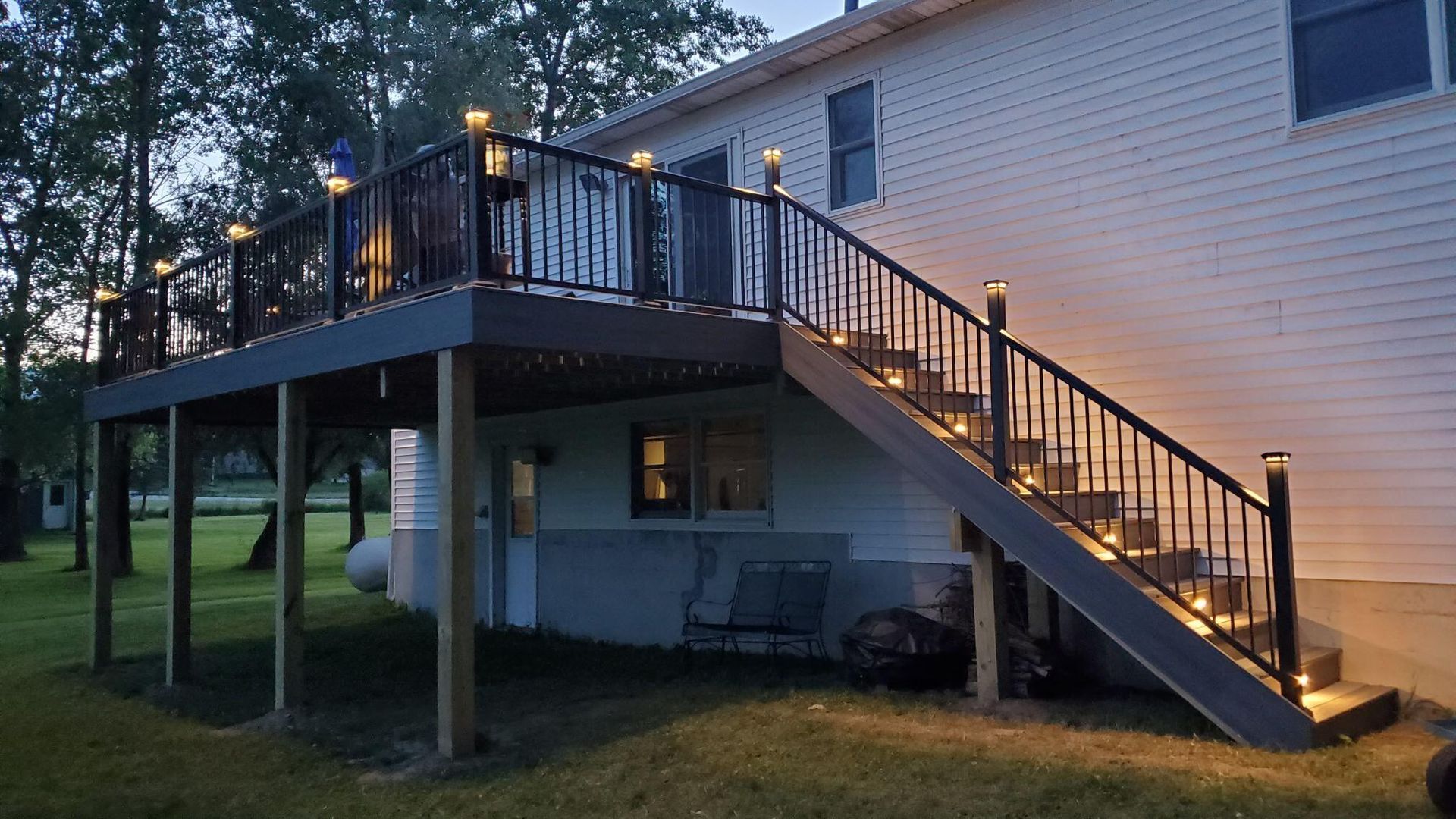 Deck with black railing and steps, lit by warm lights, attached to a two-story house.