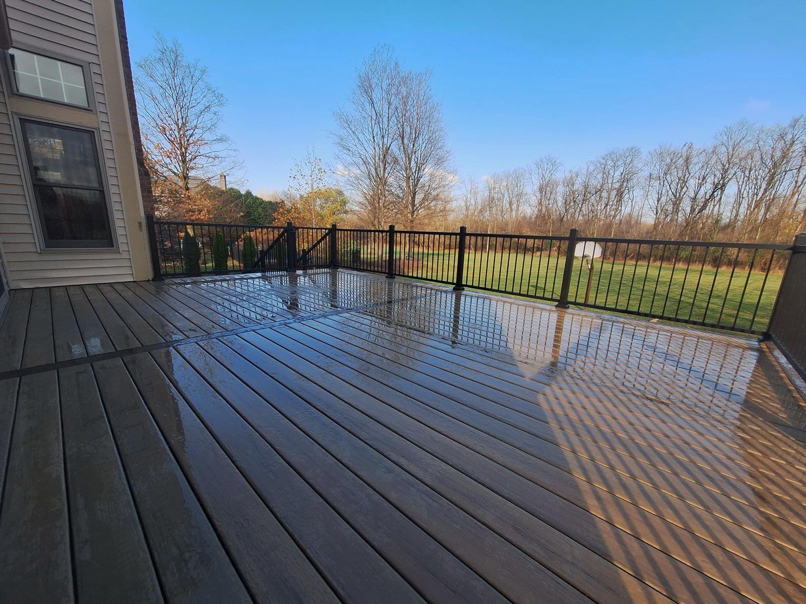 Wooden deck, wet from rain, with black railing, overlooking a grassy yard and trees under a blue sky.