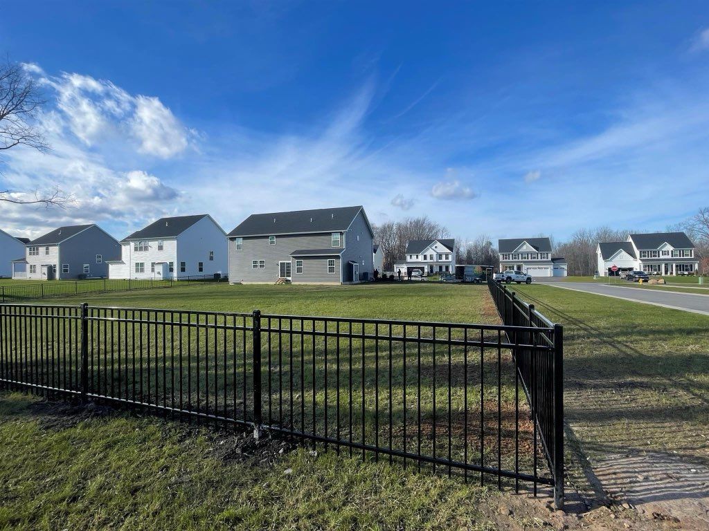 Black fence in front of grassy area, with houses in the background under a blue sky with clouds.