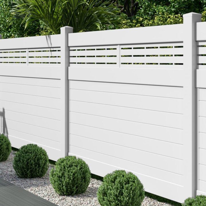 A white fence with holes in it is surrounded by bushes and gravel.