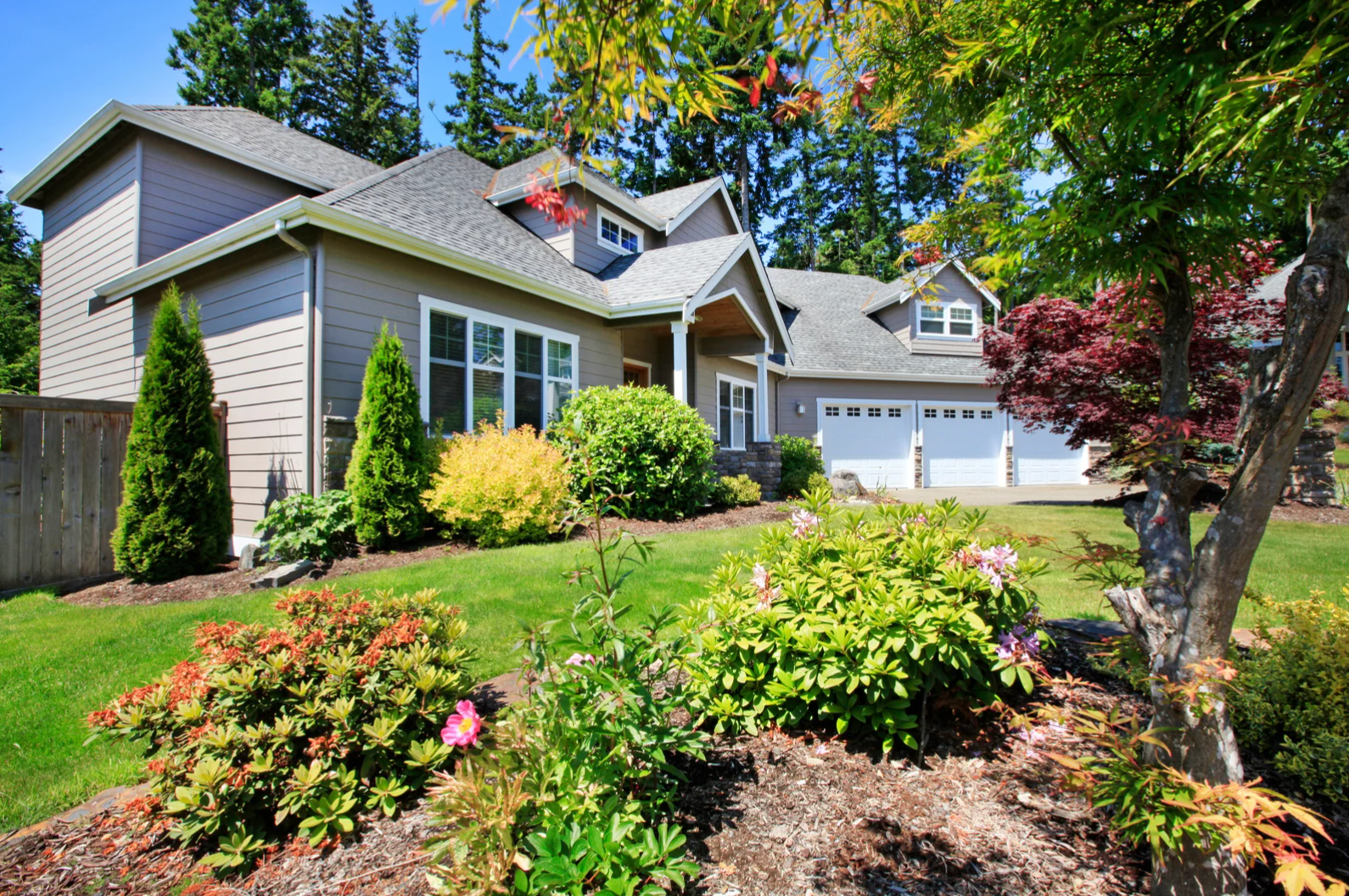 Suburban house with gray siding, green lawn, and colorful landscaping. Three-car garage and trees in the background.