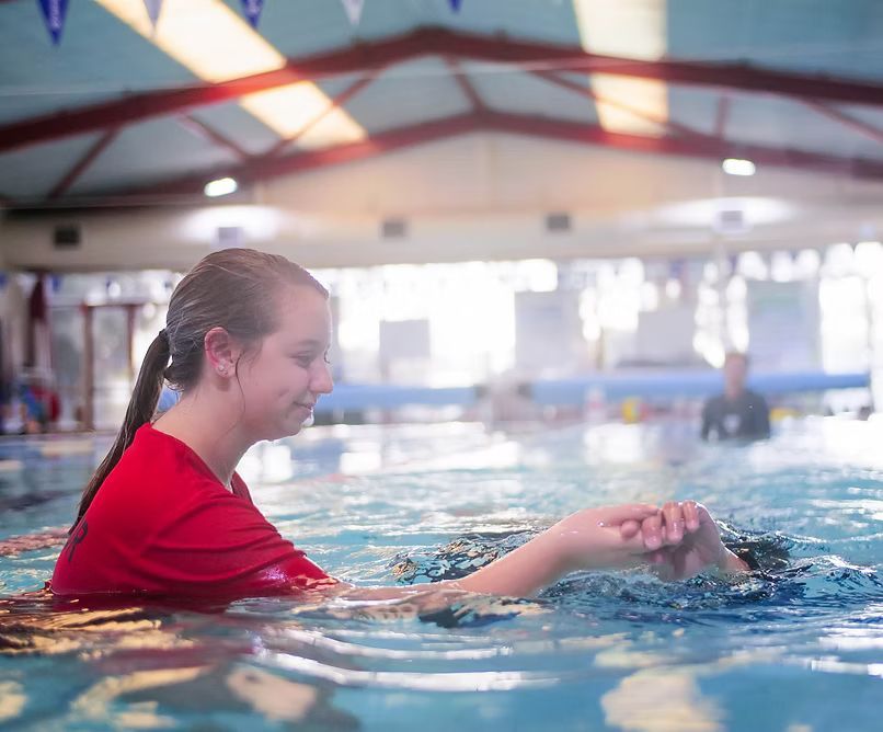 Person in red shirt in pool, holding another person's hand, near pool's edge.