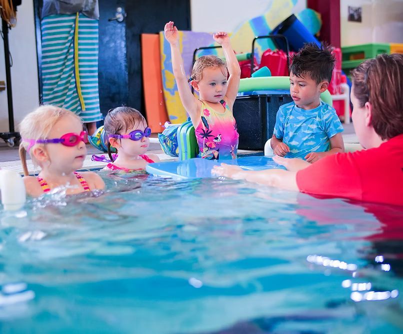 Children in a swimming pool taking a lesson, arms raised, with instructor, colorful setting.