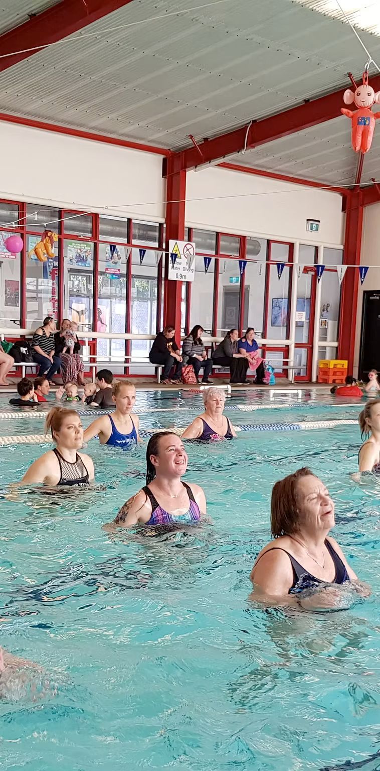 People doing water aerobics in a pool. The indoor pool has a high ceiling and red beams.